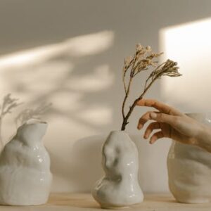 A hand gently touches a unique ceramic vase amidst a minimalist still life with soft lighting and shadows.